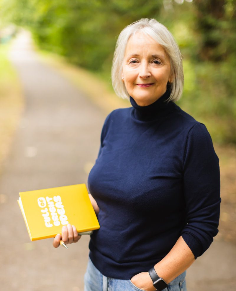 a mature woman on a towpath in a navy turtle neck holding a yellow book
